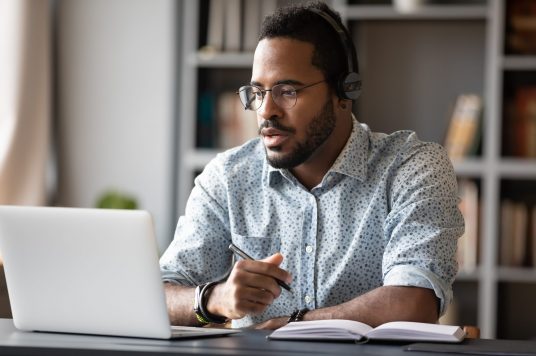 Focused african businessman wear headphones study online watching webinar Focused young african businessman wear headphones study online watching webinar podcast on laptop listening learning education course conference calling make notes sit at work desk, elearning concept